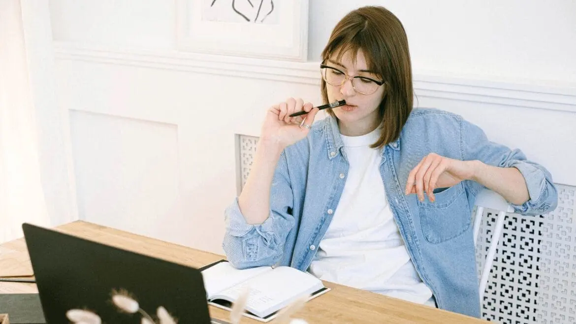 Young woman in glasses and denim shirt sitting at a desk with a laptop and notebook, holding a pen near her mouth thoughtfully.