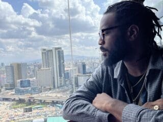 Man with sunglasses looking out a window at city skyline.