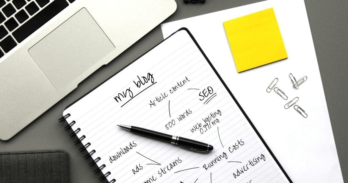 A desk setup with a laptop, a notebook titled 'My Blog' with notes on SEO and content strategies, a black pen, a yellow sticky note, and paper clips, representing blogging and content creation