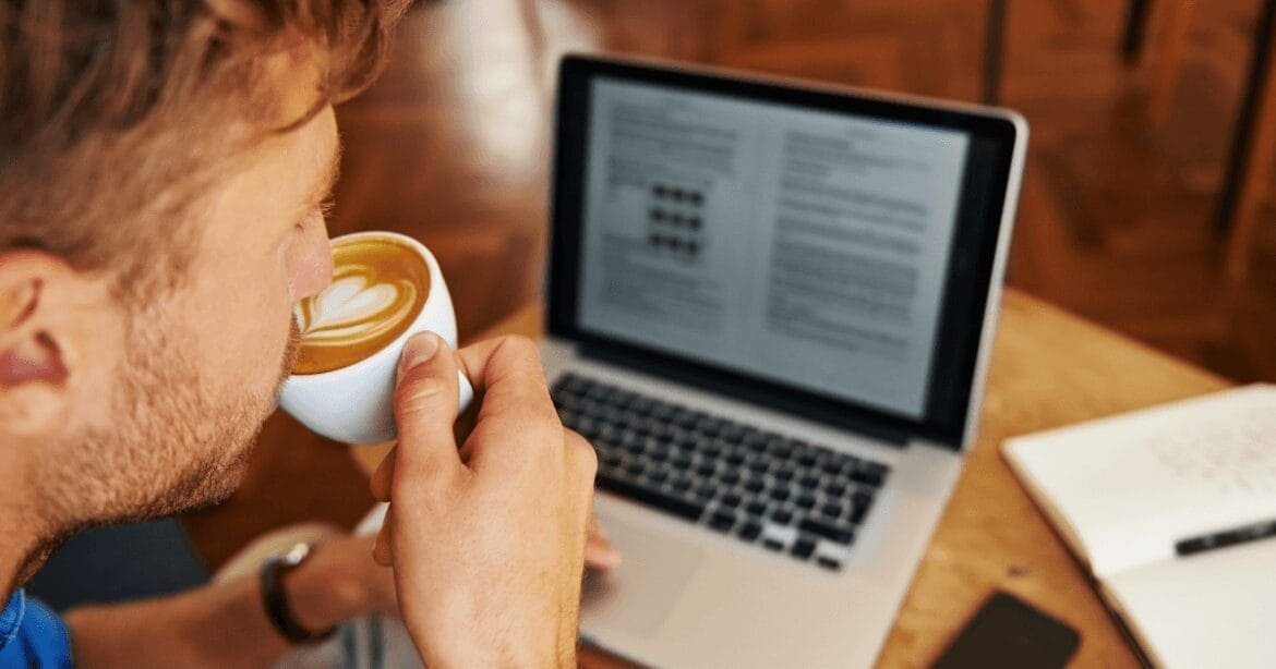 A man with light hair sips a cup of coffee while working on a laptop at a wooden table in a cozy indoor setting. An open notebook and a smartphone lie on the table beside him.