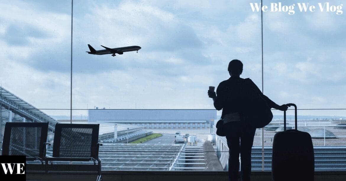 A woman standing in an airport terminal, gazing through large windows as a plane takes off into the sky, with her luggage beside her.