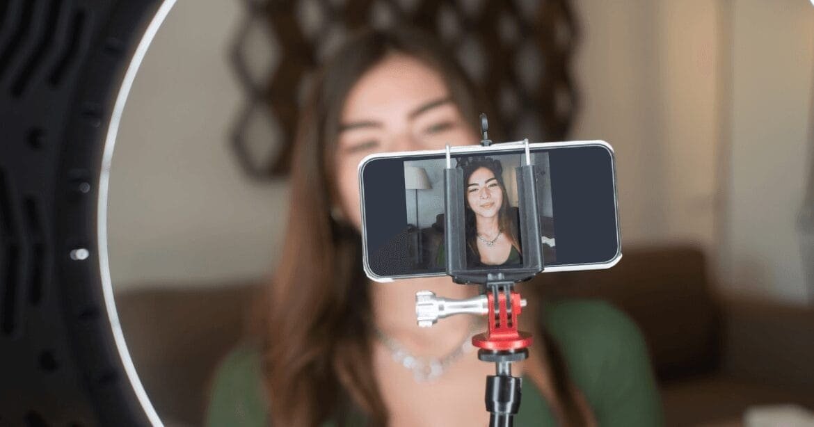 A woman filming herself using a smartphone mounted on a tripod, framed by a ring light, with a blurred background of a modern living space. The phone's screen displays her image, suggesting she is recording a video or live-streaming.successful blogging and vlogging
