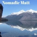 A professional portrait of Nomadic Matt, a well-known travel blogger, smiling and wearing casual outdoor clothing, with a blurred travel-themed background.