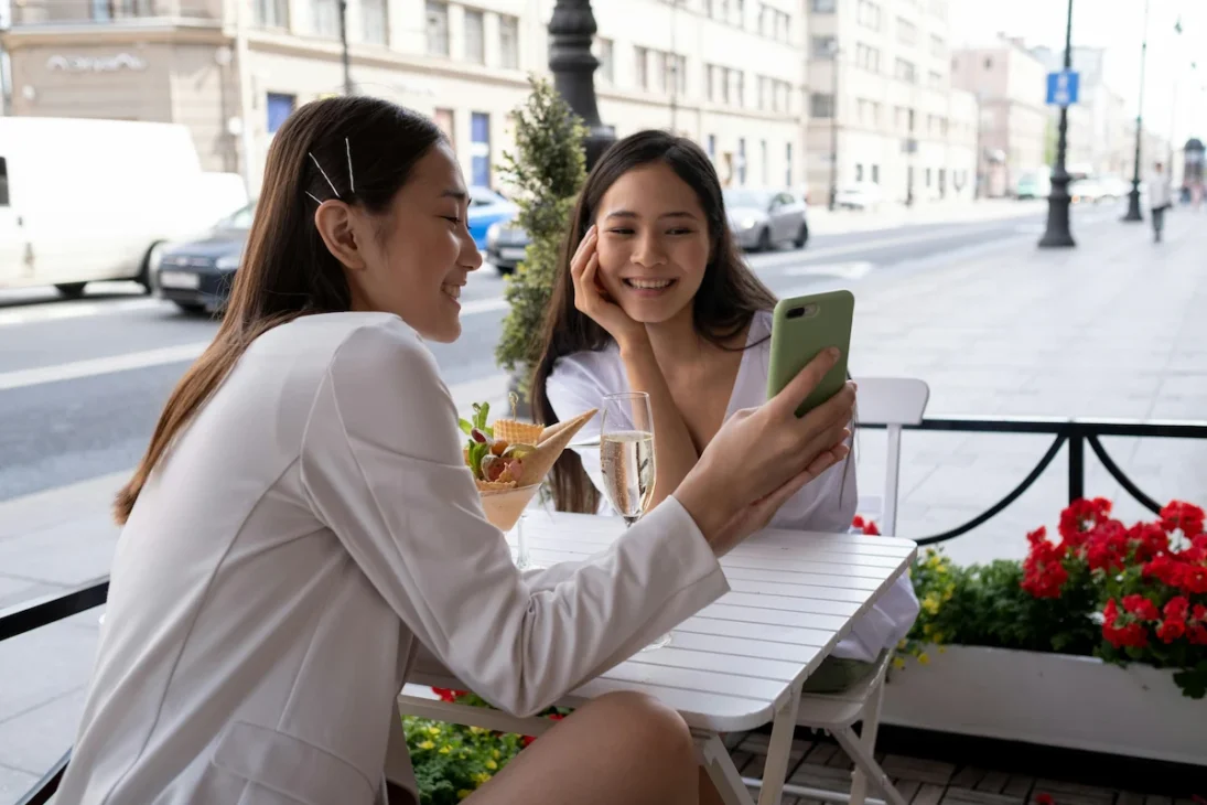 Two Gen Z girls using smartphones outdoors, laughing at outdated internet slang words like “slay” and “vibe check.”
