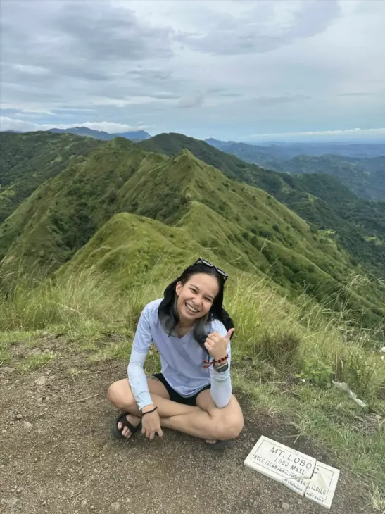 Leigh sitting on the ground at the peak of Mt. Loboc, smiling and giving a thumbs up, reflecting joy, personal goals, and a positive mindset