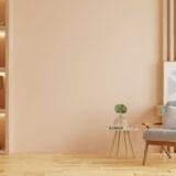 A cozy living room corner with a gray armchair, small round side table with a plant and glasses, wooden floor, built-in shelves with decorative items, and soft peach-colored walls.