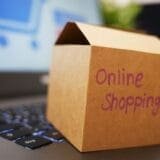 A small cardboard box labeled "Online Shopping" sits on a laptop keyboard, with a blurred shopping cart icon visible on the laptop screen in the background.