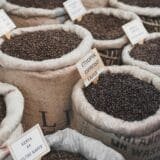 Multiple burlap sacks filled with dark roasted coffee beans are displayed in a market or store setting. Each sack has a small sign indicating the type of coffee beans, such as "Etiyopya Espresso Kahve" and "Kenya AA Filtre Kahve." The sacks are neatly arranged, showcasing the rich, glossy coffee beans.
