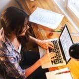 Woman typing on a laptop at a wooden desk with documents and a desk lamp nearby.