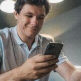 Man in a light blue polo shirt smiling as he looks at his smartphone.