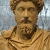 Close-up photo of a marble bust of a Roman man with a curly beard and detailed clothing, displayed in a museum setting.