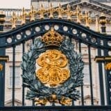 Decorative black and gold wrought iron gate with royal insignia, featuring a crown and intricate designs, in front of Buckingham Palace.