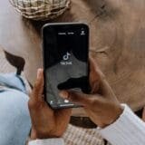 Close-up of a person holding a smartphone with the TikTok app loading on the screen. The phone is held above a wooden table, and another person sits nearby, indicating a casual social setting.