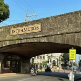 A stone archway labeled "Intramuros" marks the entrance to the historic district in Manila, with motorcycles passing underneath and a yellow "Bike Lane" sign visible on the roadside.