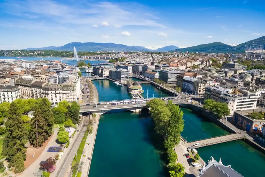 Aerial view of Geneva, Switzerland, showing Lake Geneva, the city center, and surrounding Alps under clear skies.