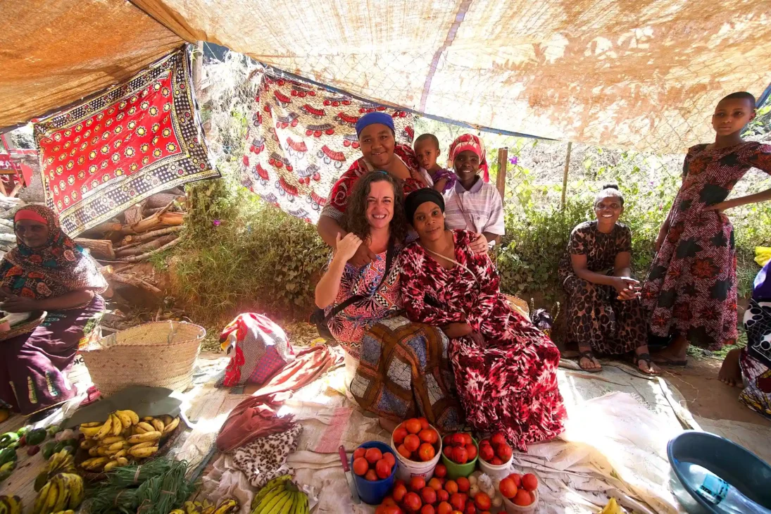 A lively market stall in Lushoto with a group of people sitting under a makeshift canopy. Bright fruits like tomatoes and bananas are arranged in buckets on the ground, surrounded by patterned textiles, trees, and locals chatting.