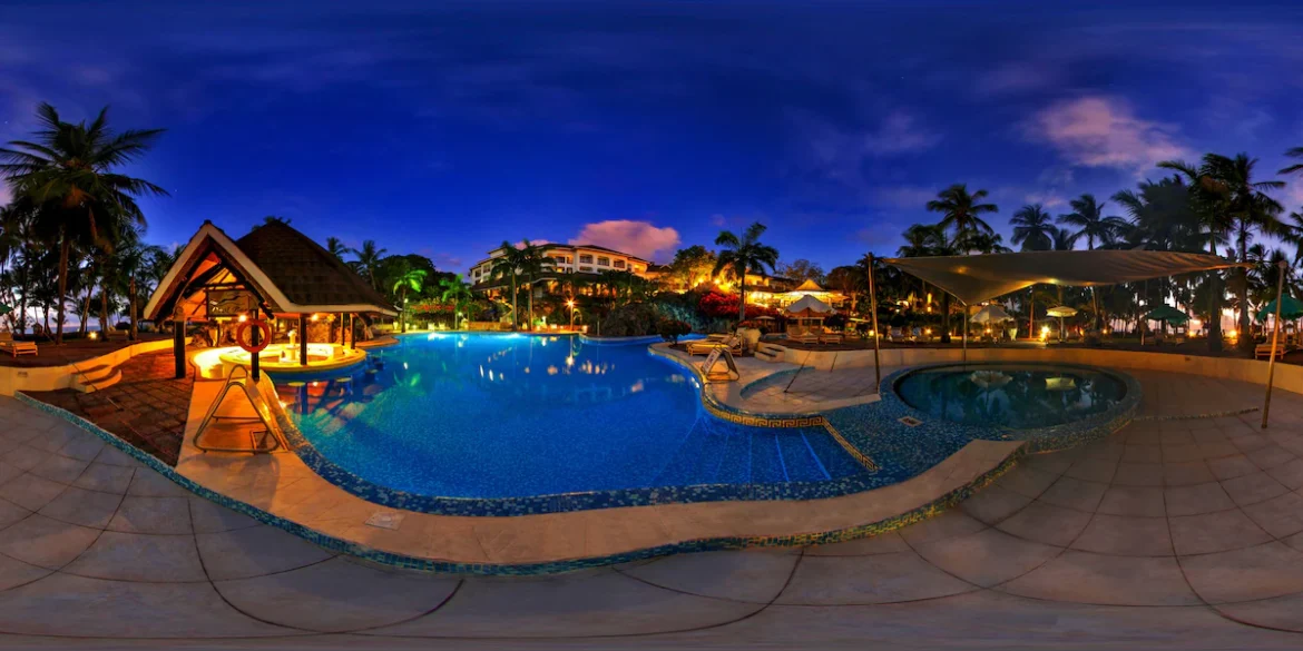 Panoramic night view of a luxurious resort swimming pool in Diani, Mombasa, surrounded by bright blue lighting, a poolside bar hut, and palm trees, highlighting the luxury side of Mombasa travel.