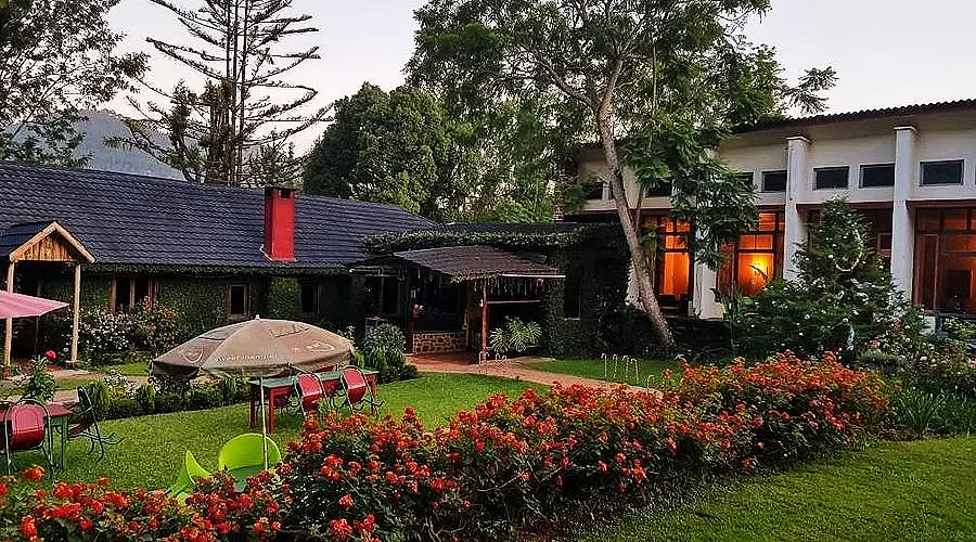 A cozy garden courtyard of a villa or hotel, with red flowerbeds lining a manicured lawn, outdoor seating under umbrellas, and a mix of rustic and modern buildings surrounded by trees.