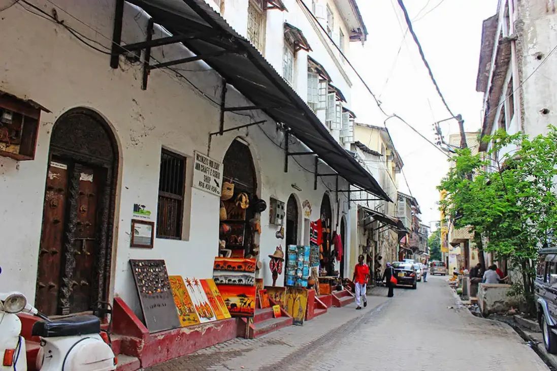 Street view of a narrow alley in Mombasa Old Town, lined with historic buildings, small shops displaying local crafts, and a parked scooter, reflecting authentic Mombasa travel.