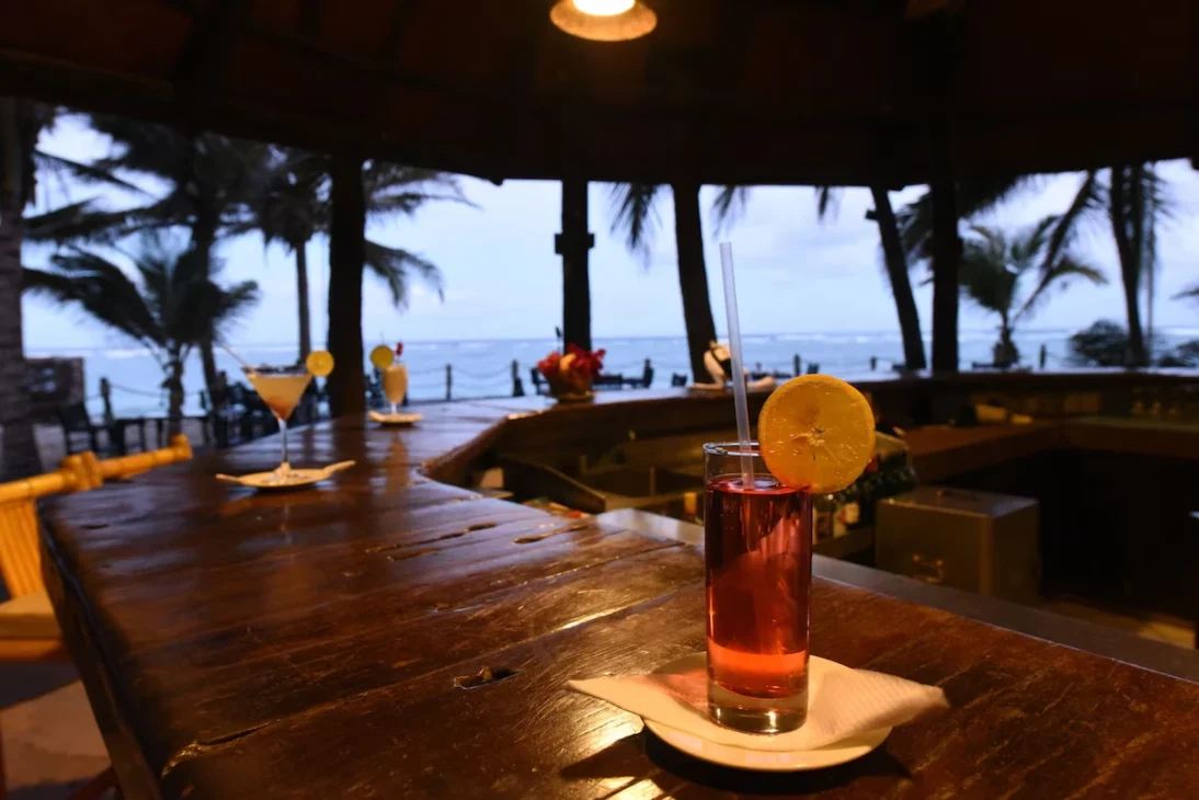 Close-up of cocktails on a wooden bar counter with palm trees and the ocean visible in the background at dusk, highlighting the relaxing side of Mombasa travel.