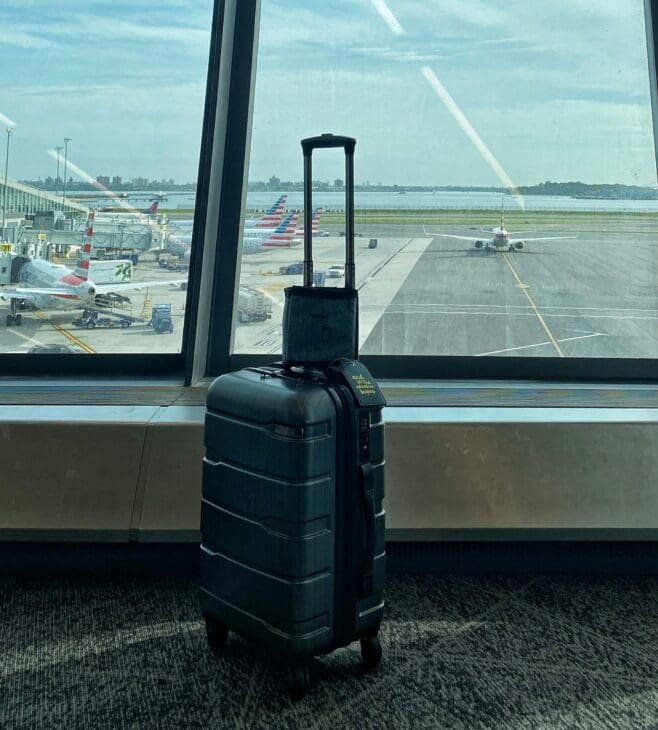 A dark-colored roller suitcase stands alone by a large airport window overlooking the tarmac with several planes parked at gates, symbolizing delayed or lost luggage.