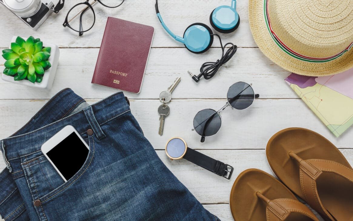 A flat lay of travel accessories including a scarf, hat, minimalist jewelry, and versatile shoes arranged neatly on a neutral background.