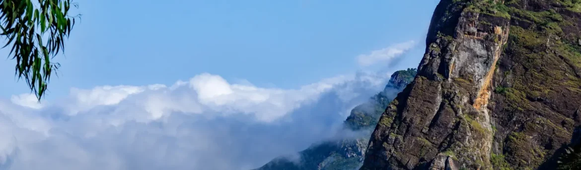 Rocky cliffside rising above low-lying clouds over a lush green hillside of Usambara Mountains, with blue sky and a hint of trees.
