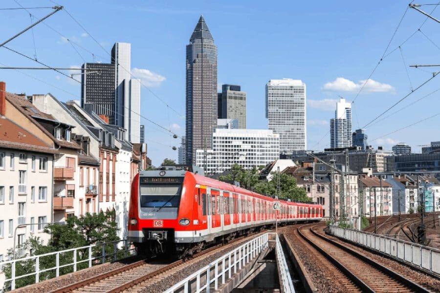 A bright red Deutsche Bahn (DB) passenger train travels along elevated tracks into a modern city skyline with glass skyscrapers under a clear blue sky. The image emphasizes the integrated, fixed nature of urban rail systems.