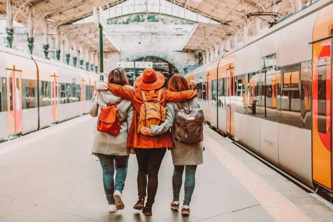 Three friends with backpacks walking arm-in-arm down a train station platform, ready to board a train. The image is taken from behind them, emphasizing their togetherness as they embark on a journey.