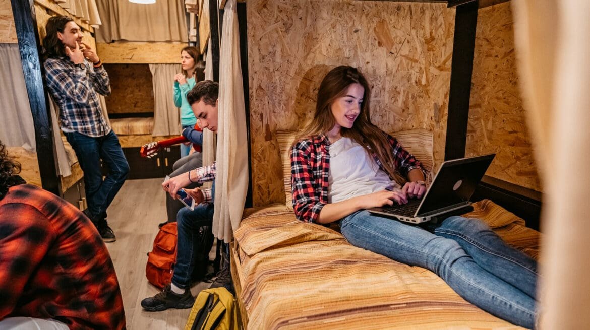 Gen Z travelers budgeting on a laptop in a hostel common area with a low-cost meal and backpack nearby, highlighting affordable travel habits.