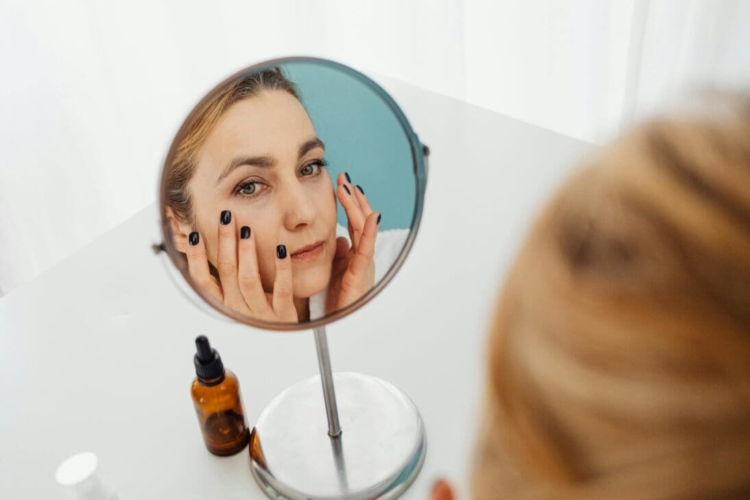 Traveler looking stressed in a hotel room mirror, examining dull and irritated skin with scattered skincare bottles on the counter.