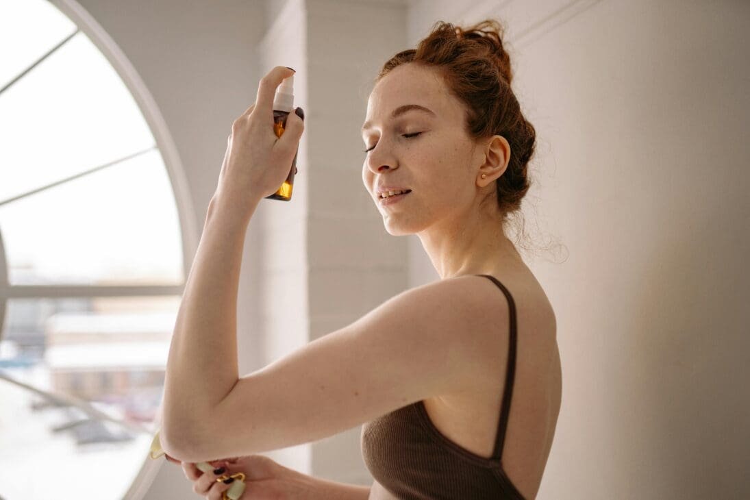 Traveler applying hydrating face mist by a sunlit hotel window, with luggage and passport nearby, showing refreshed, glowing skin.
