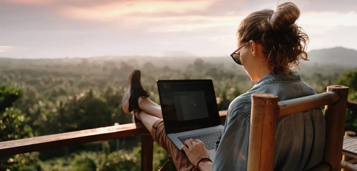 A high-angle, rear-view shot of a woman working on a laptop while sitting outdoors on a wooden balcony. She has her feet up on the railing, overlooking a lush, misty tropical forest at sunrise. She is wearing sunglasses and has her hair in a bun, embodying a relaxed "digital nomad" lifestyle in a new time zone.