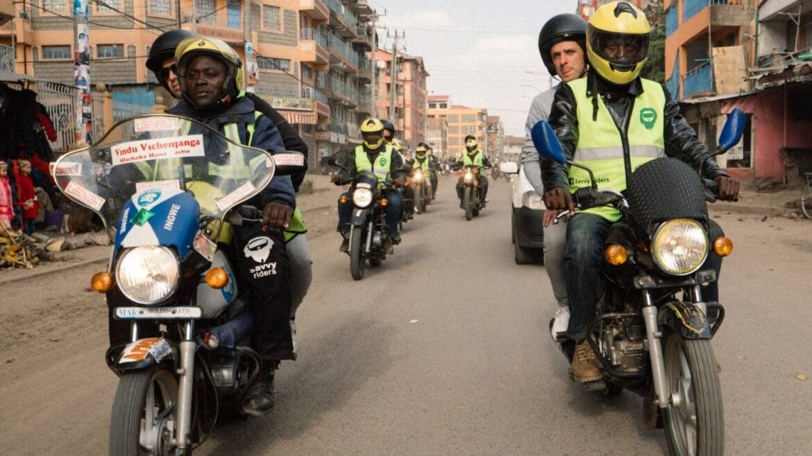 A line of boda boda motorcycle taxis driving down a Nairobi street, with riders and passengers wearing yellow safety vests and helmets.