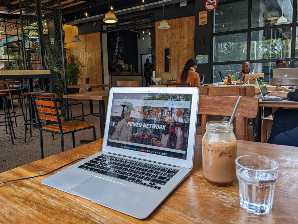 A first-person view of a remote work setup on a wooden table in a Kigali cafe, featuring a silver laptop displaying a "Power Network" website next to an iced coffee and a glass of water, with other patrons working in the background.