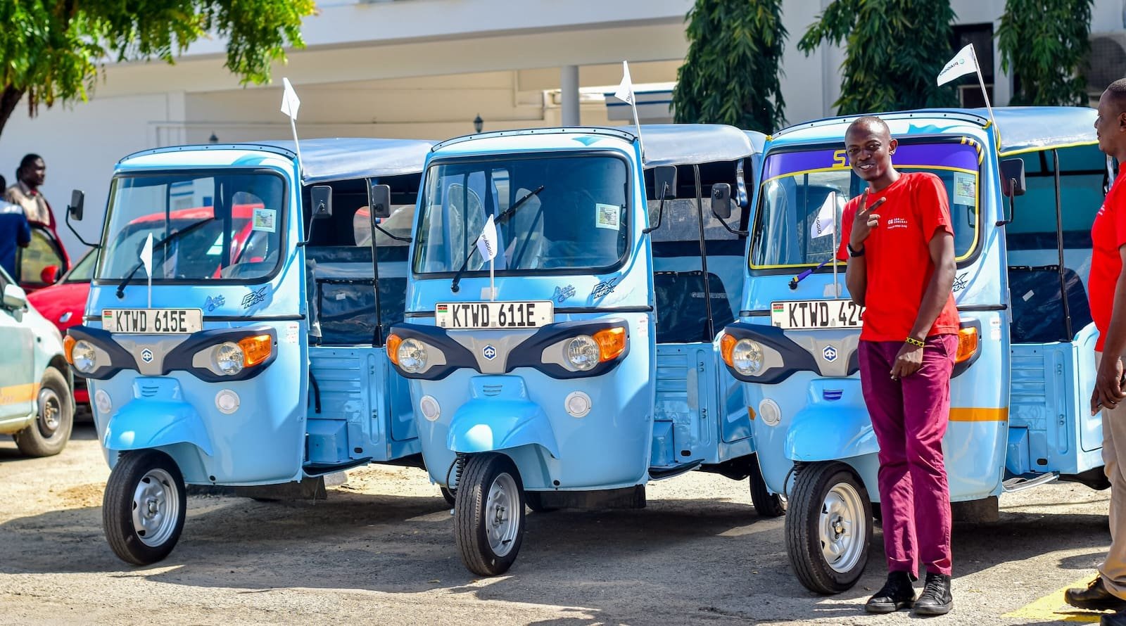 Three light blue electric tuk-tuks parked in a row in Nairobi, Kenya, with a man in a red shirt standing next to one of them smiling.