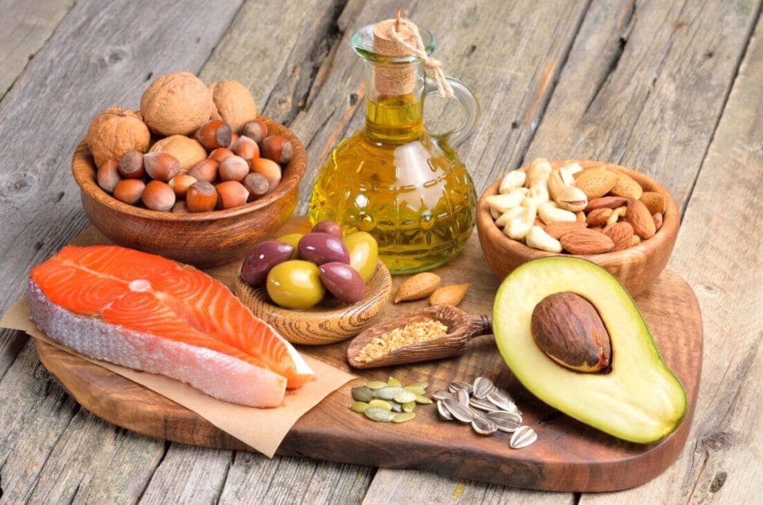 Healthy fat sources including avocado, olive oil, nuts, and salmon arranged on a wooden surface.