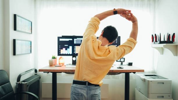 Man stretching arms and back in front of his desk and computer while working from home.