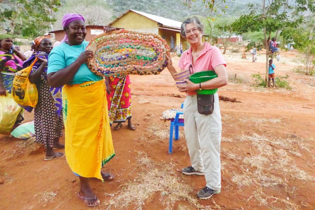 A local vendor and a tourist exchange items and laugh together in a colorful Kenyan market filled with textiles and crafts.