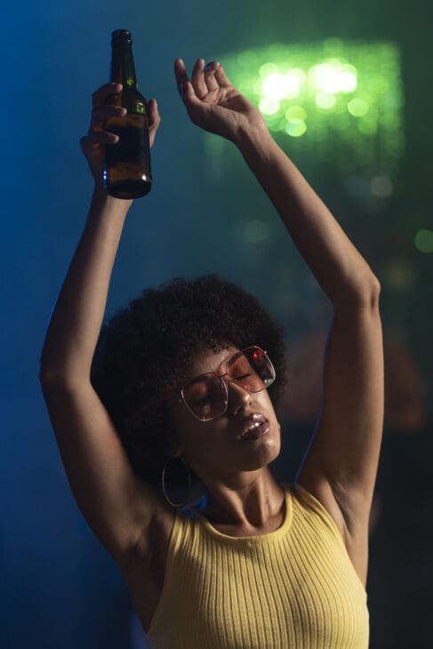 A woman with a beautiful afro and large sunglasses dances with her eyes closed and arms raised, holding a beer bottle. She is bathed in vibrant blue and green club lights, fully immersed in the music and the moment.