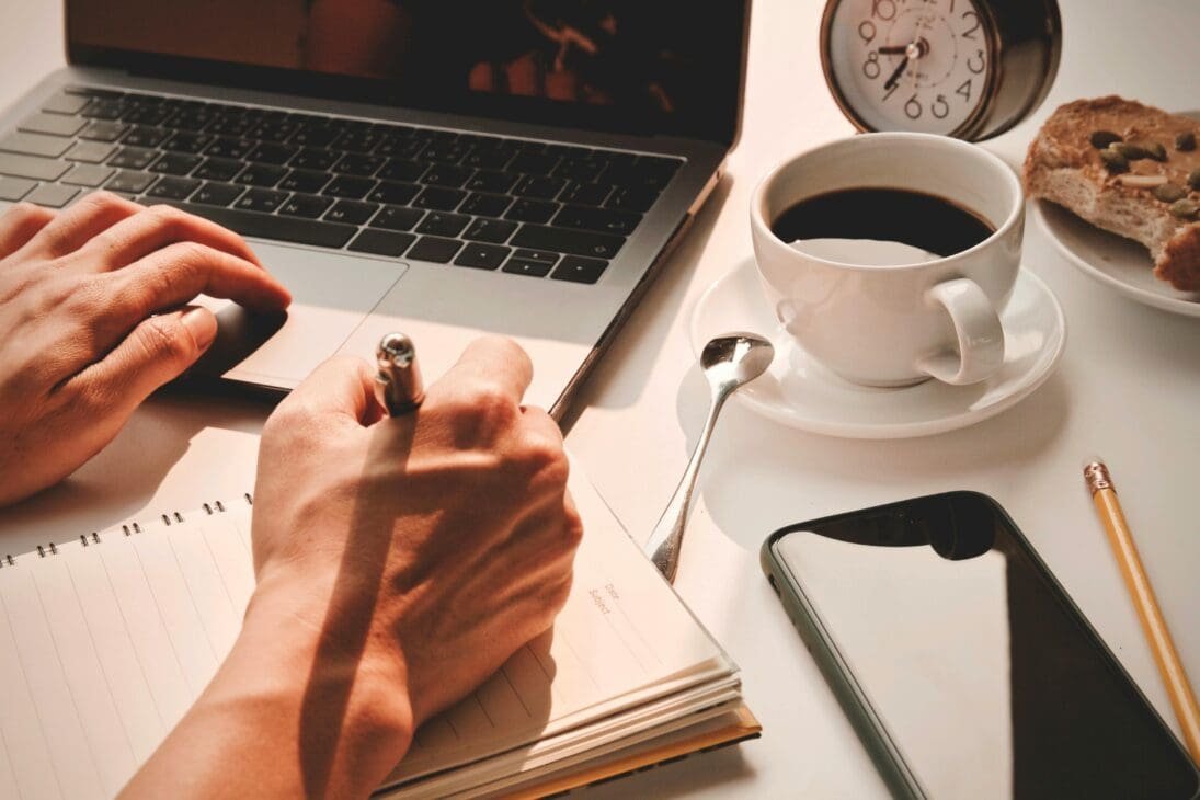 A close-up of hands writing daily goals in a planner, with a coffee cup and a small plant nearby, symbolizing routine and consistency.