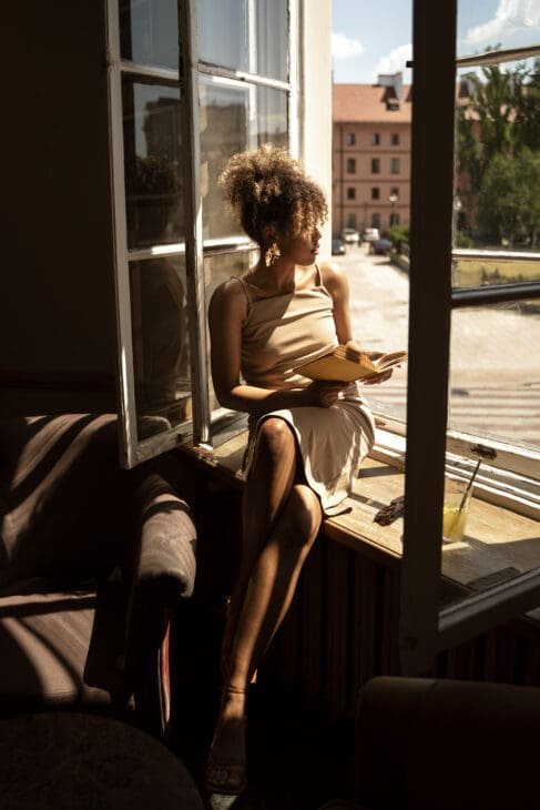 A woman in a beige dress sits on a sunny windowsill, holding an open book and gazing out an open window at a bright city street.
