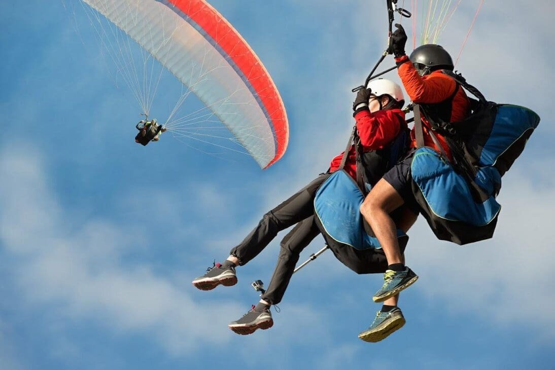 A person with a helmet and red jacket is tandem paragliding with an instructor against a blue sky with some clouds, with another paraglider visible in the distance.