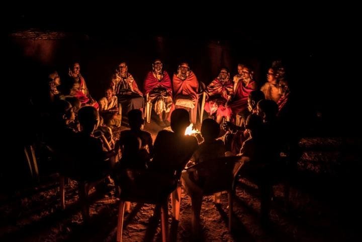 A group of people seated around a campfire in the dark; silhouettes and warm glow highlight traditional Maasai clothing and a shared moment of storytelling.