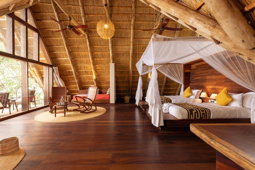 The spacious, warm wooden interior of a luxury banda at Nile Safari Lodge, featuring two king-sized beds with mosquito nets, hand-carved rocking chairs, and a floor-to-ceiling glass wall overlooking the lush riverbank.