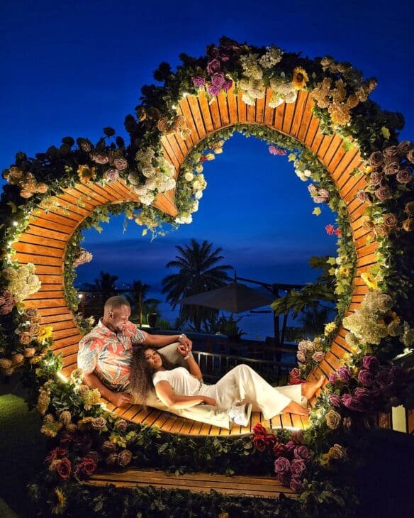 A large wooden heart-shaped frame decorated with red and pink roses overlooking a luxury resort pool and Lake Victoria at sunset.
