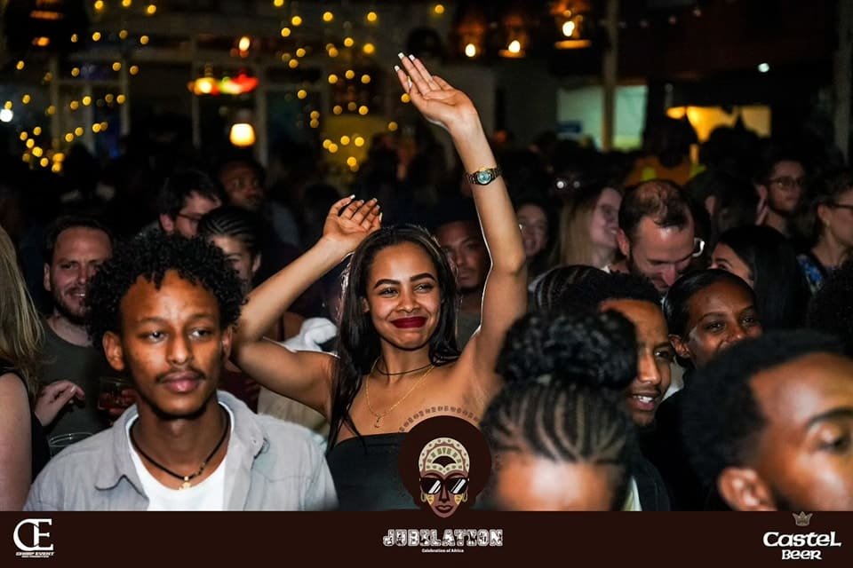 A group of stylish young Ethiopians celebrating at the Jubilation event in Addis Ababa. A smiling woman in a black top has her hands raised in the air, surrounded by a vibrant crowd under warm string lights, featuring the Jubilation and Castel Beer logos.