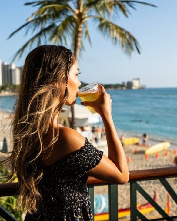 A young woman with long hair stands on a balcony overlooking a sunny tropical beach in the Philippines, drinking from a glass while looking out at the turquoise ocean and palm trees during a serene morning.