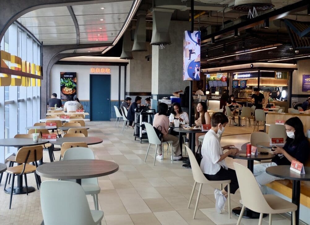 Busy mall atrium with families, friends, and couples gathering, eating, and socializing in a safe, air-conditioned environment.