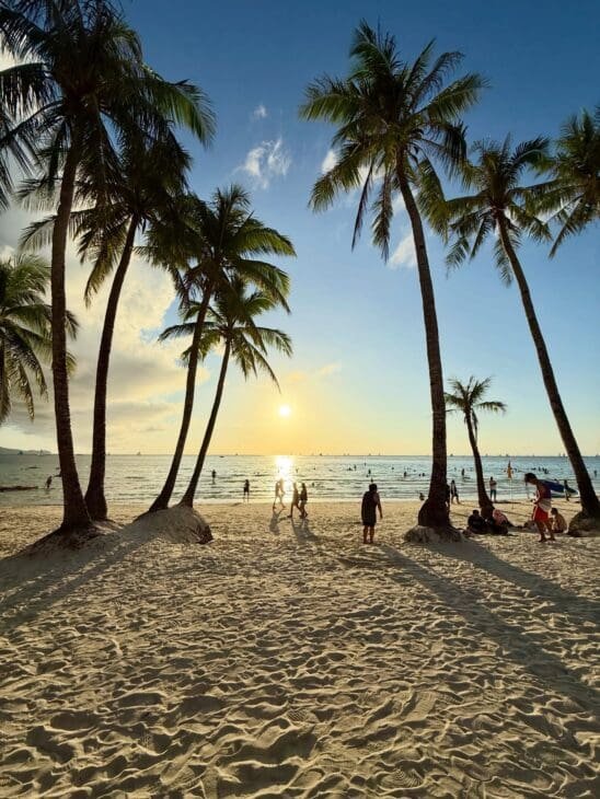 A serene tropical beach in the Philippines at sunset, featuring tall palm tree silhouettes against a golden sky, white sand with textured footprints, and people peacefully enjoying the calm ocean waters in the background.